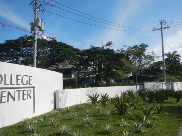 College Center-Eingangsschild an einer Wand umgeben von Grün, einem Weg, Strommasten, Bäumen, Gebäuden, einem Kran und einem bewölkten Himmel.