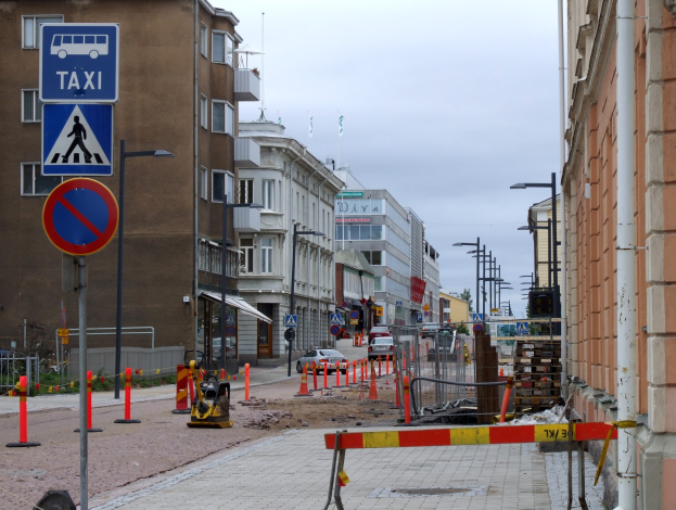 Eine Stadtstraße mit Gebäuden, Straßenlaternen, Verkehrsschildern, Absperrpollern, Bäumen und einer Baustelle mit Verkehrsschildern unter einem bewölkten Himmel.