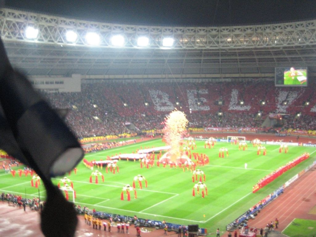 Ein Stadion mit Flutlicht, Eisengrills, Zuschauern, stehenden Personen, Werbetafeln, Anzeigetafeln, Sicherheitskamera, Flaggen und Dekorationselementen.
