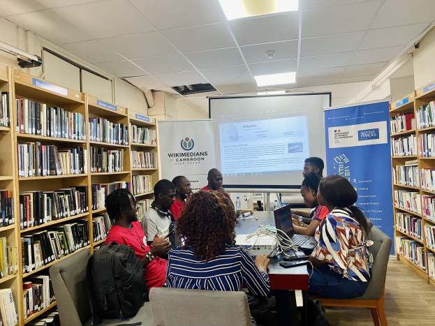 Gruppe von Menschen, die um einen Tisch in einer Bibliothek sitzen, mit Laptops, Flaschen und Taschen, die in eine Diskussion vertieft sind, in der Nähe von Bücherregalen, Projektionsbildschirm und Schildern mit Text.