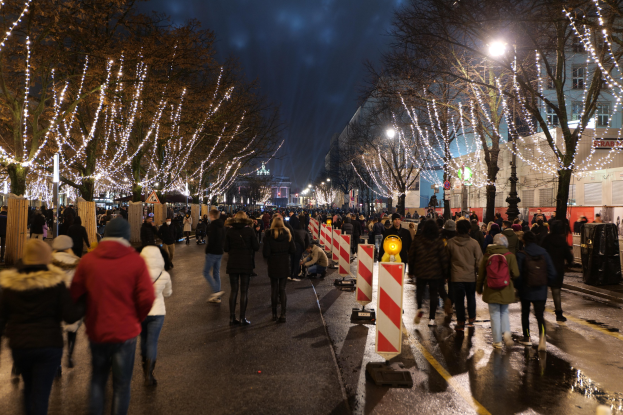 Eine Menschenmenge, die nachts eine Straße entlanggeht, beleuchtet von festlichen Weihnachtslichtern, mit Bäumen und Gebäuden im Hintergrund unter einem bewölkten Himmel.
