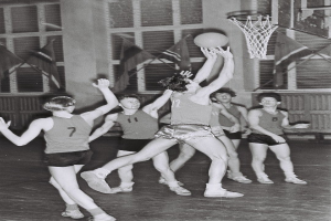 Schwarzes und weißes Foto von jungen Männern, die Basketball in einer Turnhalle spielen, mit einem Basketballkorb in der oberen rechten Ecke und Flaggen im Hintergrund.