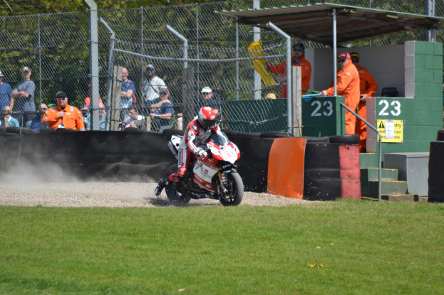 Ein Motorradfahrer mit Helm und Handschuhen fährt über ein grasbewachsenes Feld mit Zuschauern in Kappen und Brillen, Absperrungen, Treppen mit Geländern und einem nummerierten Schuppen im Hintergrund.
