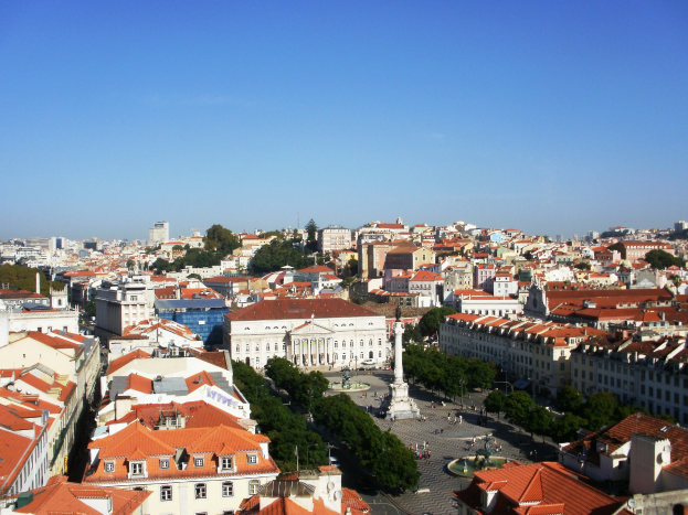 Blick auf Lissabon von einem Hügel mit Gebäuden mit Fenstern, Bäumen, einer Statue auf einem Sockel, wenigen Menschen auf der Straße und dem Himmel im Hintergrund.