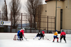 Menschen, die Eishockey auf einem Eisplatz mit Gebäuden, Bäumen, einer Straßenlaterne, einer Namensplakette und Zäunen im Hintergrund unter einem Himmel spielen.