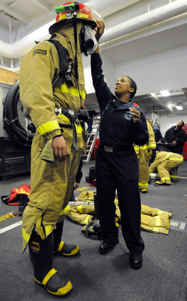 Zwei Feuerwehrleute, eine in einem schwarzen Kleid und Helm und ein anderer in gelb-schwarzer Ausrüstung, stehen in der Nähe einer Leiter und anderer Ausrüstung mit Menschen im Hintergrund.