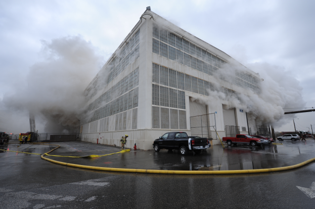 Ein großes Industriegebäude steht in Flammen mit Rauch, der herausquillt, umgeben von Fahrzeugen und einigen Menschen, mit einem wolkenverhangenen Himmel im Hintergrund und einem Wasserzeichen auf dem Bild.