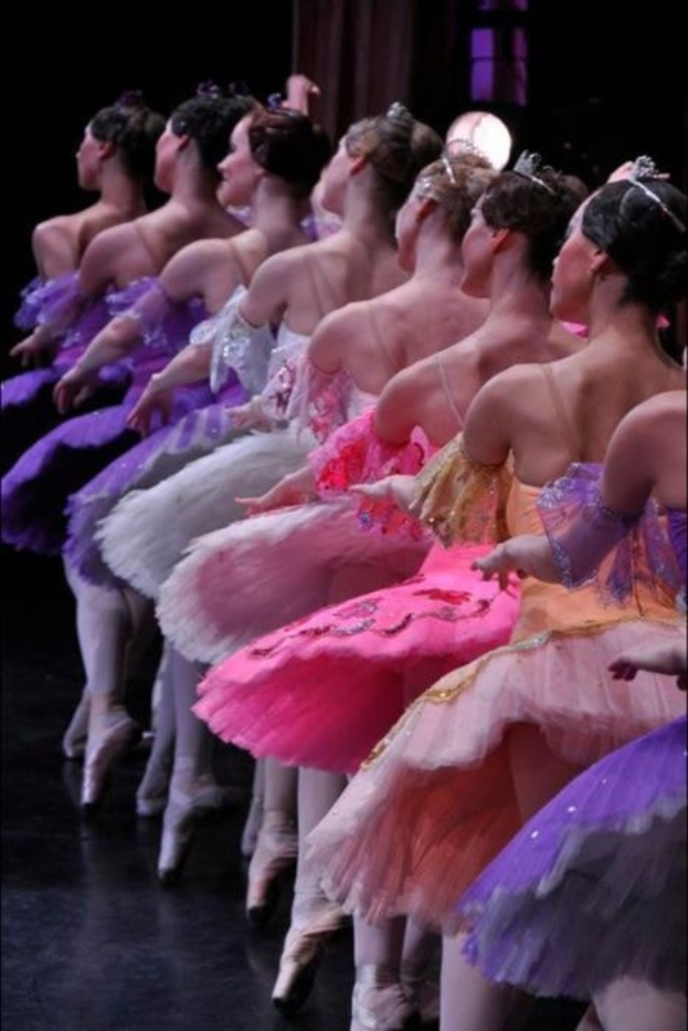 Eine Gruppe von Balletttänzerinnen in rosafarbenen Tutus sitzt auf einer Bühne, beleuchtet von einem Licht im Hintergrund, ihre synchronen Bewegungen schaffen eine fesselnde Szenerie.
