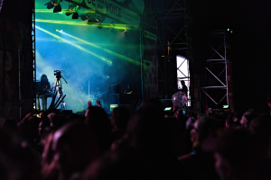 Ein volles Konzert mit Musikern auf einer hell erleuchteten Bühne, mit Instrumenten, Mikrofonen und einem Banner vor einem dunklen Hintergrund.
