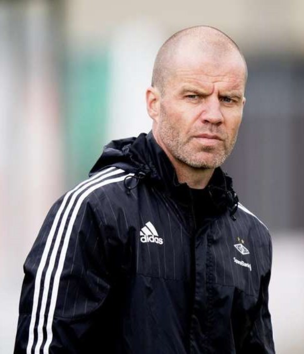 Bald Trainer in einer blauen Jacke mit weißen Streifen und weißem Kragen auf einem Fußballfeld mit gefalteten Händen und ernster Miene.