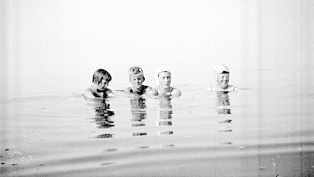 Schwarzes und weißes Foto von vier Kindern, die in einem Pool schwimmen und alle Schwimmkappen tragen.