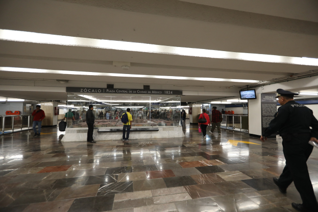 Ein überfüllter U-Bahnhof mit einem Polizeibeamten im Vordergrund, Menschen mit Taschen, Überhangtexttafeln und Lichter, sowie Geländer im gesamten Bereich.