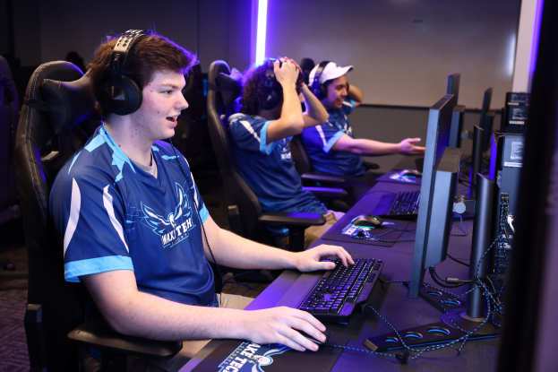 Eine Gruppe junger Männer trägt Headsets und spielt ein Videospiel an Computern auf einem Schreibtisch mit Monitoren, Tastaturen und anderen Gegenständen, während hinter ihnen Lichter an der Wand zu sehen sind.