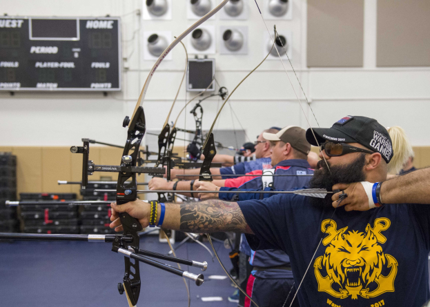 Eine Gruppe von Bogenschützen in Mützen beim Indoor-Training während der World Archery Championships 2017, mit einer Texttafel links und Ausrüstung rechts.
