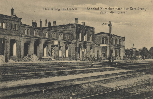 Altes Schwarz-Weiß-Foto des Bahnhofs Korschen in Ostern, Deutschland, zeigt einen Bahnhof mit Bahnschienen, Gebäuden, Strommasten, Menschen, Bäumen und einem Himmel im Hintergrund.