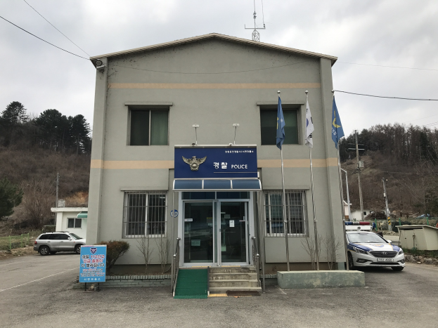 Außenansicht des Seoul Police Headquarters-Gebäudes mit Glasfenstern, Treppenaufgang, Fahnenmasten, parkenden Fahrzeugen, Infotafel, Bäumen, Strommasten und bewölktem Himmel.