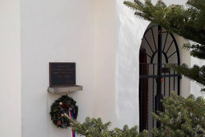Ein weißes Gebäude mit einem Kranz, einer Gedenktafel und einem Baum auf der rechten Seite.