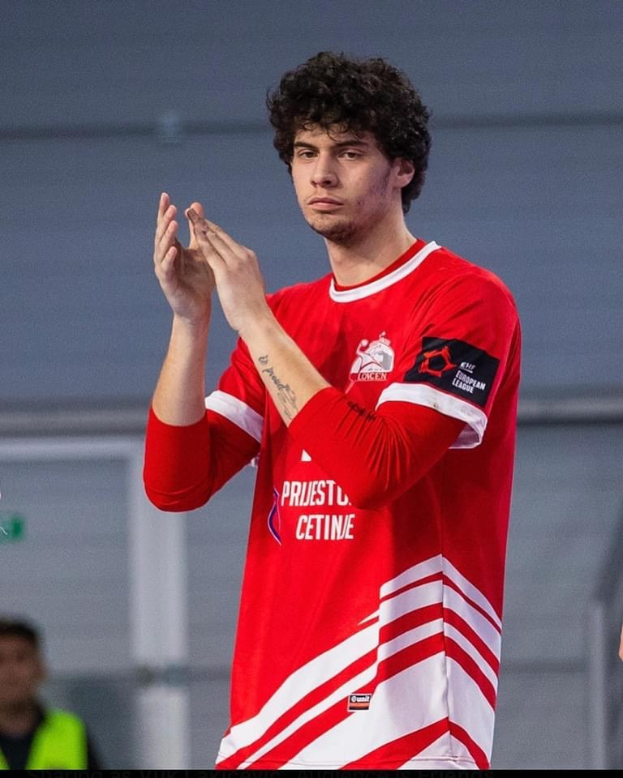 Ein Mann in einem roten und weißen Hemd klatscht in den Händen in der Vordergrund, mit einer anderen Person und einer Wand im Hintergrund und Text, der unten 'Kopenhagen Futsal Club - Foto 1' liest.