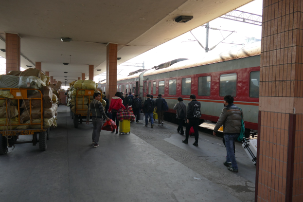 Menschen gehen auf einem Bahnsteig an einem stehenden Zug vorbei, mit Gepäckwagen auf der linken Seite und Säulen auf der rechten Seite, unter Stromleitungen gegen einen klaren blauen Himmel.