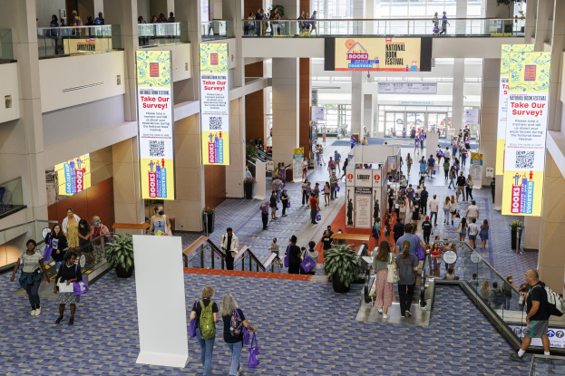 Große Gruppe von Menschen, die durch ein großes Gebäude mit Bannern, Topfpflanzen, Rolltreppen, Geländern, Säulen, Texttafeln, Glastüren und anderen Gegenständen geht, einige tragen Taschen, wahrscheinlich an der Nationalen Buchmesse teilnehmend.