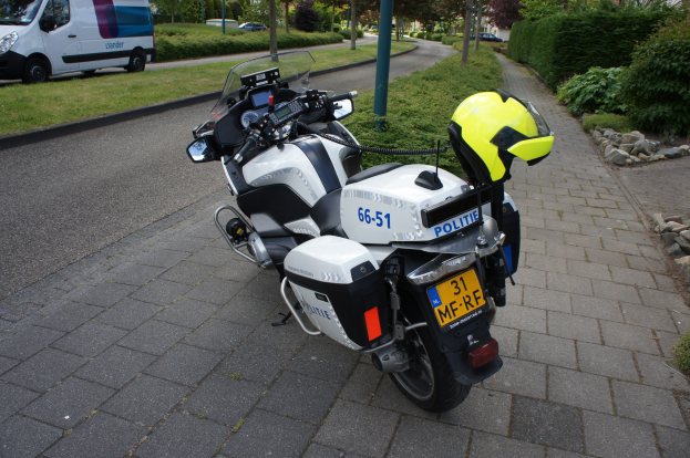 Ein auf der Seite der Stra√e geparktes Polizeimotorrad mit einem Helm darauf, umgeben von B√∞umen, Masten, Gebäuden, Pflanzen, Gras, Steinen und anderen Fahrzeugen im Hintergrund.
