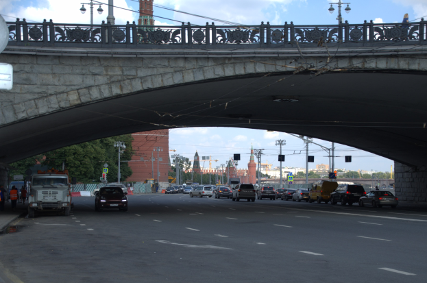 Eine Brücke überspannt eine vielbefahrene Straße mit darunter fahrenden Fahrzeugen, während Fußgänger auf dem Gehweg links stehen, mit städtischen Elementen wie Verkehrsampeln, Bäumen, Gebäuden und Laternen im Hintergrund.