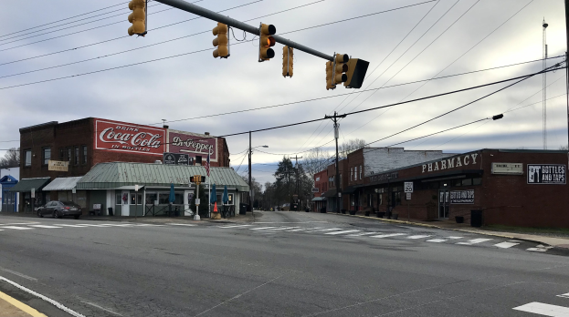 Stadtstraßenkreuzung mit einer Ampel, Fahrzeugen, Gebäuden mit Beschriftung, Strommasten mit Kabeln, Bäumen und einem bewölkten Himmel.