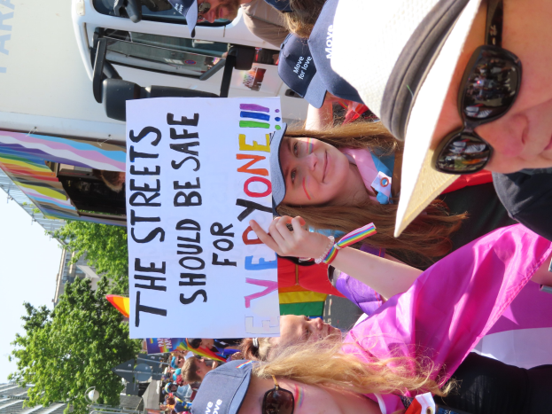 Eine Gruppe von Menschen hält ein "Die Straßen sollten sicher sein"-Schild vor einem Lastwagen, einige tragen Mützen und Schutzbrillen, andere halten Schilder, vor einem Gebäude, Bäumen und einem klaren blauen Himmel.