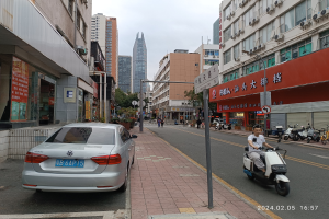 Ein Mann fährt mit einem Roller die Straße zwischen hohen Gebäuden entlang, mit Menschen auf den Gehwegen und Fahrzeugen auf der Straße, unter einem klaren blauen Himmel mit Bäumen im Hintergrund.