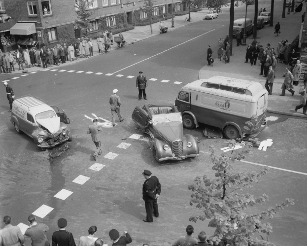 Schwarz-weiß-Szene eines Autounfalls an einer Straßenkreuzung mit mehreren Fahrzeugen, darunter ein Van, und einer Gruppe von Schaulustigen; Gebäude, Bäume, Strommasten und Texttafeln sind im Hintergrund sichtbar.