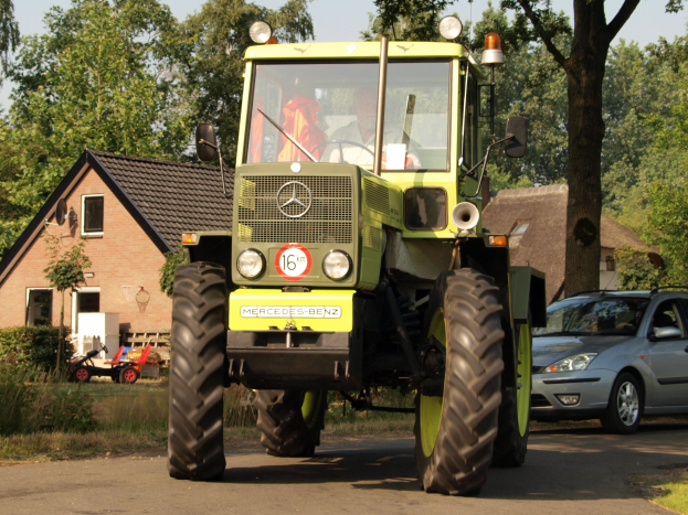 Grüner Traktor fährt auf einer Straße neben einem Auto, mit einer Person auf dem Fahrersitz des Traktors, Häusern und Bäumen im Hintergrund unter einem klaren blauen Himmel.