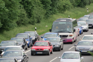 Verkehrsstau auf einer Autobahn mit vielen Autos und einem Lieferwagen, Menschen in den Fahrzeugen, mit Bäumen und Gras im Hintergrund.