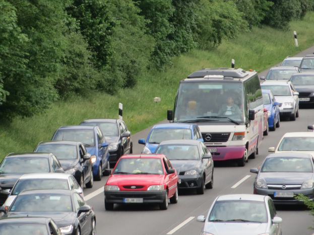 Verkehrsstau auf einer Autobahn mit vielen Autos und einem Lieferwagen, Menschen in den Fahrzeugen, mit Bäumen und Gras im Hintergrund.