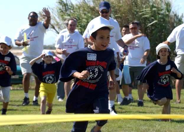Kinder laufen in einem Wettbewerb auf einem grünen Rasen mit einem gelben Band im Vordergrund, einige tragen Kappen und Schutzbrillen, andere halten Gegenstände, mit Bäumen und einem klaren blauen Himmel im Hintergrund.