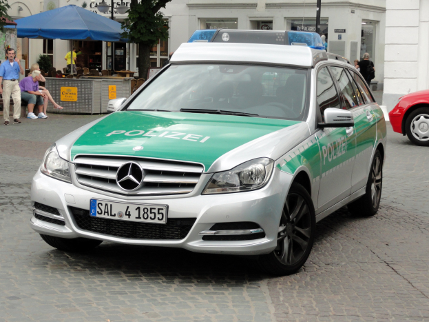 Ein schwarzer Mercedes-Benz C-Klasse W204 Polizeiwagen neben einem roten Auto auf einer Straße geparkt, mit Gebäuden, Bäumen und verschiedenen städtischen Objekten im Hintergrund.