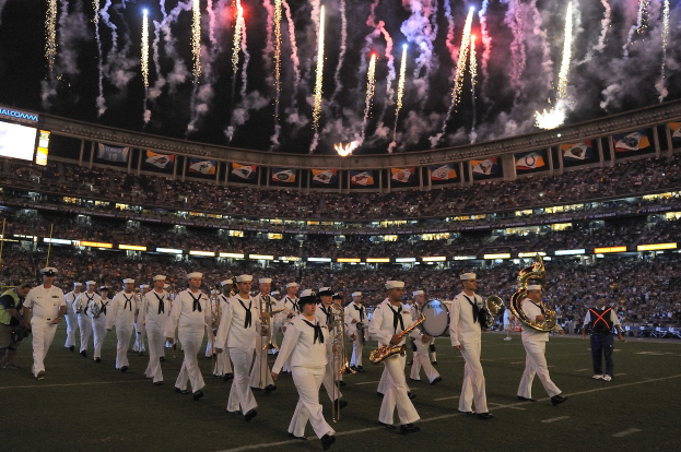 Eine Gruppe von Menschen in weißen Uniformen marschiert auf einem Fußballfeld, einige halten Musikinstrumente, während sie von einer Menge im Hintergrund mit Bannern, Bildschirmen auf der linken Seite und Feuerwerk am Himmel beobachtet werden.