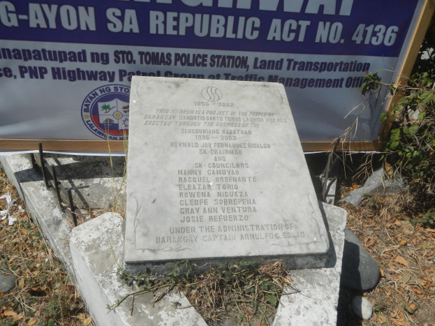 Gedenkstein mit eingravierter Inschrift, umgeben von Steinen, trockenen Blättern, Pflanzen und einer Fahne mit Text und Logo, mit einem Schild im Hintergrund, auf dem "G-Ayon SA Republic Act No. 4136" steht.