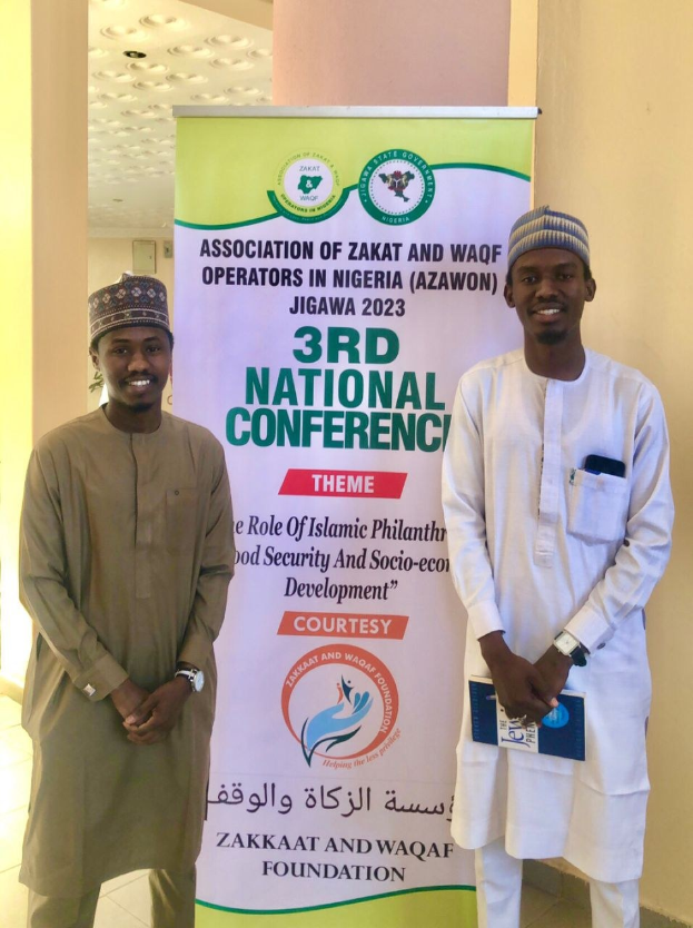 Zwei Männer stehen nebeneinander vor einem Banner mit der Aufschrift "3. Nationalkonferenz" mit einer Säule und Deckenleuchten im Hintergrund.