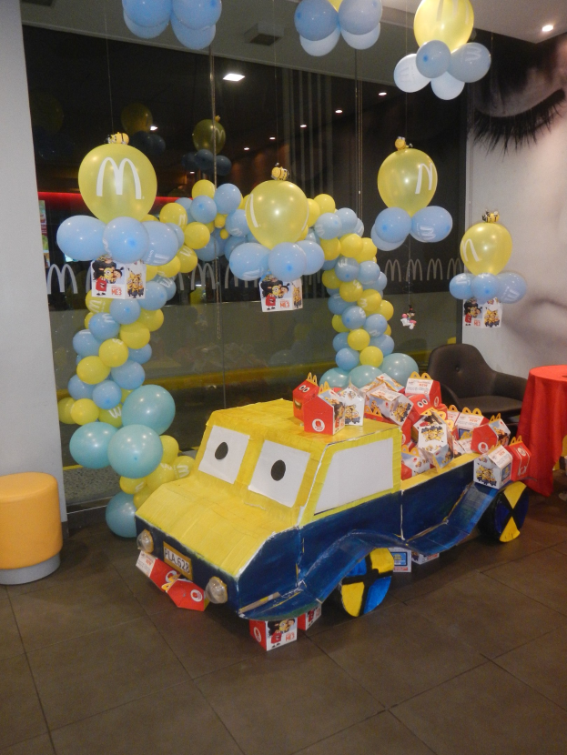 Eine festliche McDonald's-Geburtstagsfeier-Szene mit einem gelben und blauen Lastwagen, der mit Ballons geschmückt ist, einem rot gedeckten Tisch mit Gegenständen, Hockern, einer Glaswand und Deckenlampen.