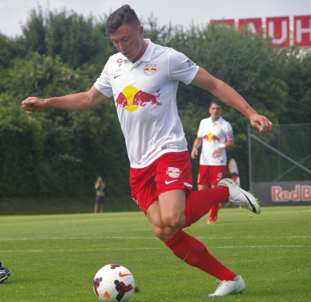 Ein professioneller Fußballspieler schießt den Ball auf einem Rasenfeld mit Bäumen und einem klaren blauen Himmel im Hintergrund, in der Nähe eines Schildes mit der Aufschrift "RB Leipzig."