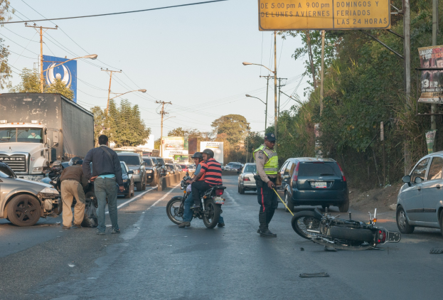 Eine Gruppe von Menschen steht um ein verunglücktes Motorrad auf der Seite der Straße herum, mit mehreren Fahrzeugen, Bäumen, Polen, Lampen, Schildern und dem Himmel im Hintergrund.