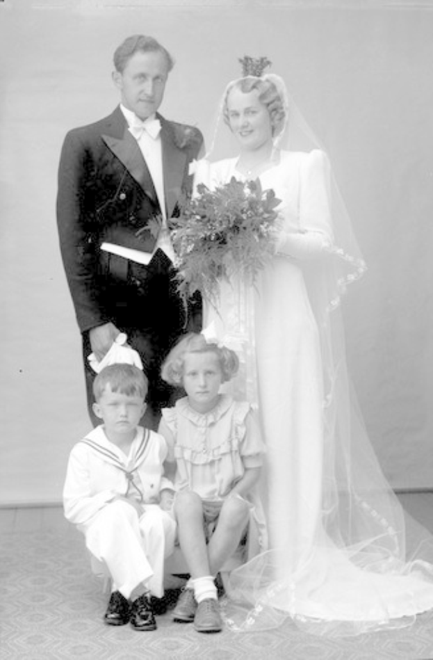 Schwarzes und weißes Foto einer Braut in einem weißen Kleid mit einem Strauß, einem grinsenden Bräutigam daneben und zwei Kindern, die vor ihnen auf dem Boden sitzen.