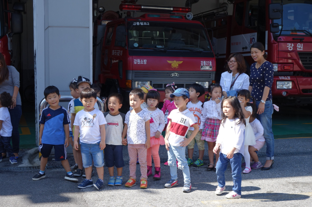 Gruppe von Kindern vor einem Feuerwehrauto an einer Feuerwache, einige mit Mötzen, mit weiteren Feuerwehrautos im Hintergrund.