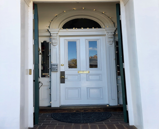 Ein weißes Haus mit einer blauen Haustür, einem Türvorleger und dekorativen Lichtern um die Tür.