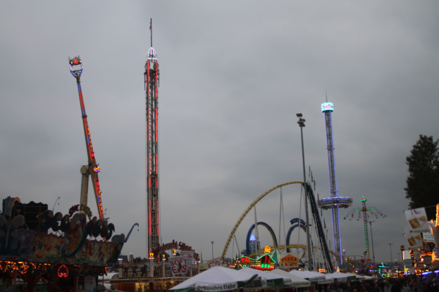 Eine Menschenmenge versammelt sich um einen nächtlichen Karneval, beleuchtet von den Lichtern der Fahrgeschäfte, Pfähle und Bäume, unter einem bewölkten Himmel.