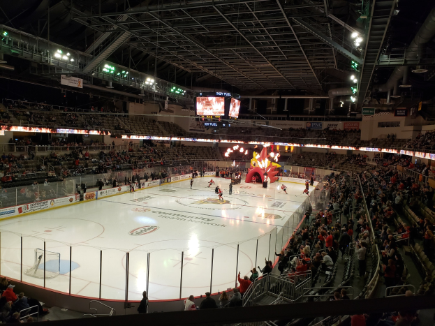 Eishockeyspiel in einer großen Arena mit Zuschauern auf Sitzen und Stehplätzen in der Nähe der Eisfläche, umgeben von Bannern und einem Dach mit Metallstangen, Lampen und zwei Bildschirmen, identifiziert als Nationwide Arena.