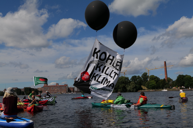 Gruppe von Menschen in Kajaks, die auf dem Wasser paddeln, mit einem Banner, das "Kohle Kill Klima" trägt, und Bäumen, Gebäuden, Kränen und einem klaren blauen Himmel im Hintergrund.