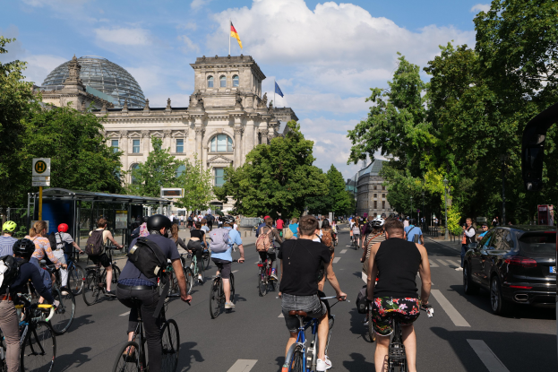 Gruppe von Menschen, die auf Fahrrädern eine von Bäumen gesäumte Straße in Berlin, Deutschland, entlangfahren, mit einer Bushaltestelle auf der rechten Seite und einer Flagge, die auf einem Gebäude unter einem bewölkten Himmel weht.