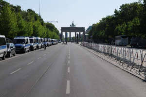 Eine Reihe von Polizeiwagen, die auf der Seite einer Straße vor dem Brandenburger Tor in Berlin, Deutschland, geparkt sind, mit Menschen, die Fahrräder fahren und auf der Straße stehen, Absperrungen und Bäume, die die Seiten säumen, und ein Tor mit Statuen im Hintergrund.