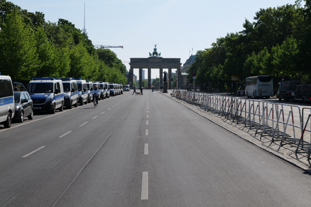 Eine Reihe von Polizeiwagen, die auf der Seite einer Straße vor dem Brandenburger Tor in Berlin, Deutschland, geparkt sind, mit Menschen, die Fahrräder fahren und auf der Straße stehen, Absperrungen und Bäume, die die Seiten säumen, und ein Tor mit Statuen im Hintergrund.